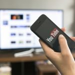 Foto: 1 de 1 Pessoa segurando um celular com logo do YouTube, aparecendo televis