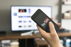 Foto: 1 de 1 Pessoa segurando um celular com logo do YouTube, aparecendo televis