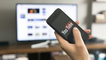 Foto: 1 de 1 Pessoa segurando um celular com logo do YouTube, aparecendo televis