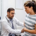 Patient telling physician about her pain and health problems during visit to hospital. Young woman complaining about back or kidney ache while sitting on examination bed at the doctor's office